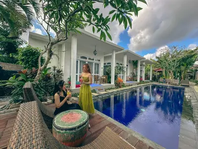 two women sitting next to a pool in front of a house at Bali Stuti Jimbaran Inn in Jimbaran
