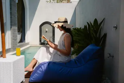 a woman sitting on a pillow reading a book next to a pool at Dikubu Bali Villa and Suites in Kerobokan