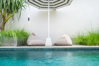 a pool with pillows next to a white wall and an umbrella at Kin House Bali in Seminyak