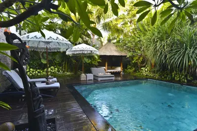 a pool in a garden with chairs and an umbrella at Elok Villas in Seminyak