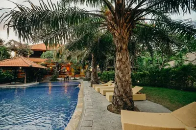 a swimming pool with a palm tree and some chairs at Three Brothers Bali Legian Bungalows & Villas in Legian